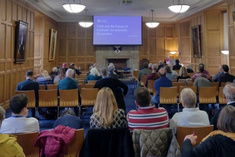 The back of a group of people sitting in a wood panelled room with paintings on the wall. They are all facing a screen that displays the writing 'Faith and the Future of Scotland: An Interfaith Perspective